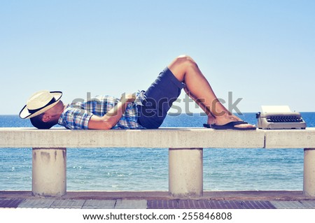 an old typewriter with a blank page and a young man lying down in a street bench near the sea, while is waiting for inspiration