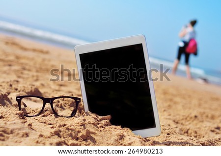 a pair of black plastic-rimmed eyeglasses and a tablet computer with a black blank space in the screen, in the sand of a beach, and a blurred woman in the background