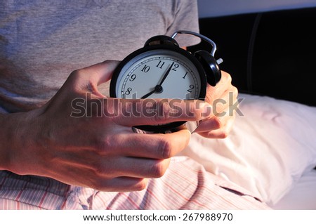 a young caucasian man wearing pajamas in bed setting the alarm clock at 7 before lie down