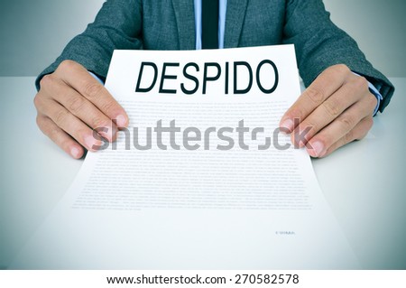 a young caucasian businessman in grey suit sitting at his office desk shows a document with the text despido, dismissal in spanish, written in it
