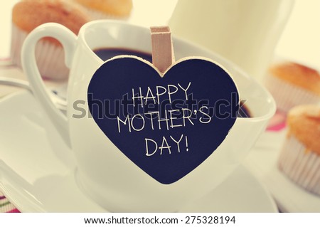the sentence happy mothers day written in a heart-shaped blackboard placed in a cup of coffee, with some muffins in the background in a set table for breakfast