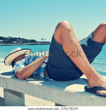 a young caucasian man with a straw hat on his face lying down in a street bench near the sea