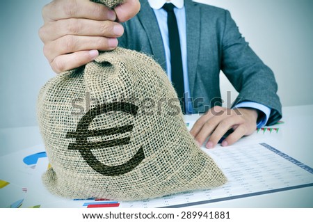 a young man wearing a gray suit seated at an office desk full of charts and financial balances holds a burlap money bag with the euro currency sign in his hand