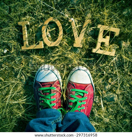 closeup of the feet of a man wearing red sneakers stepping on the grass where a pile of wooden letters form the word love, with a filter effect