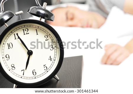 closeup of an alarm clock at 6.55 in the morning on the night table and a young caucasian man sleeping in bed