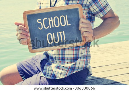 a young caucasian man sitting in a wooden pier shows a chalkboard with the text schools out written in it, with a filter effect