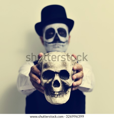 a young man with calaveras makeup, wearing top hat, holds a scary skull in front of him