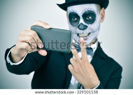 a young man with calaveras makeup, wearing bow tie and top hat, takes a selfie of himself with a smartphone