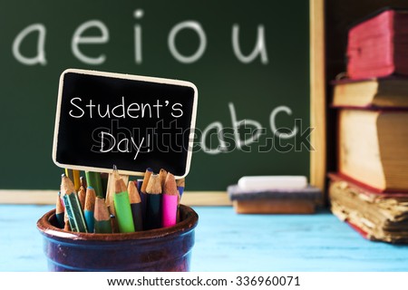 closeup of a pot with some pencil crayons and a chalkboard with the text students day written in it, placed on a blue wooden school desk, with some books, an eraser and a piece of chalk