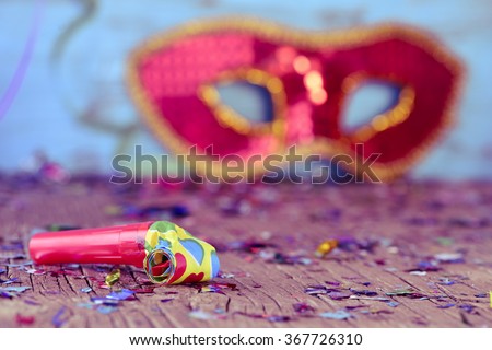 closeup of a party horn on a rustic wooden surface full of confetti and an elegant red and golden carnival mask in the background