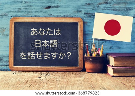 a chalkboard with the question do you speak Japanese? written in Japanese, a pot with pencils, some books and the flag of Japan, on a wooden desk