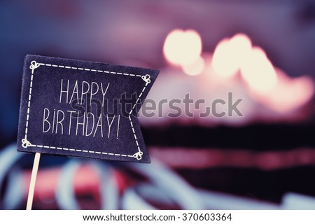 closeup of a black flag-shaped signboard with the text happy birthday, and a birthday cake with lit candles in the background