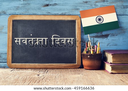 a chalkboard with the text Independence Day written in Hindi and a flag of India, on a rustic wooden surface, against a blue wooden background