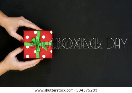 high-angle shot of a young caucasian woman with a gift in her hands wrapped in a red paper patterned with white dots and tied with a green ribbon, and text boxing day written on a dark gray surface