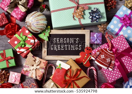 high-angle shot of some christmas ornaments, cookies, a pile of gifts tied with ribbons of different colors, and a chalkboard with the text seasons greetings written in it, on a rustic wooden surface