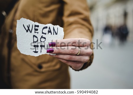 closeup of a young caucasian woman in the street showing a piece of paper with the text we can do it written in it
