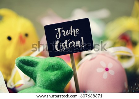 a chalkboard with the text frohe ostern, happy easter in german written in it and a pile of decorated easter eggs of different colors, a green rabbit and a yellow teddy chick in the background