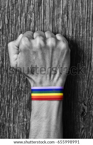 closeup of a young caucasian man in black and white with a colorful band patterned as the rainbow flag tied to his wrist and his fist raised against a rustic wooden background