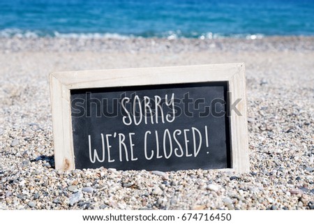 a wooden-framed chalkboard with the text sorry we are closed written in it, placed on the sand of a beach, with the sea in the background