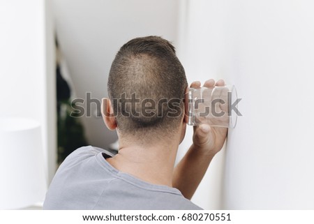 closeup of a young caucasian man seen from behind trying to hear through the wall with a glass
