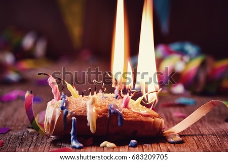 a small cake topped with some lit candles and some other unlit candles after blowing out the cake, on a rustic wooden table, sprinkled with confetti and a colorful garland in the background