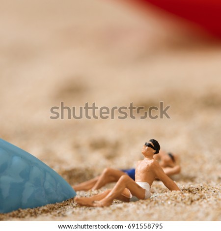 miniature man and miniature woman wearing swimsuit relaxing next to a blue plastic starfish on the sand of the beach