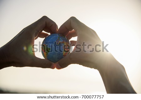 closeup of a young caucasian man with a world globe in his hands forming a heart against the sky and a ray of sun showing up on the right