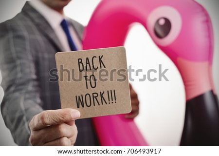 closeup of a young caucasian businessman wearing a swim ring in the shape of a pink flamingo showing a brown signboard with the text back to work written in it