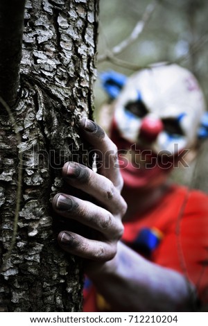 closeup of a scary evil clown wearing a dirty red costume with a creepy smile in his face while is hiding behind a tree in the woods