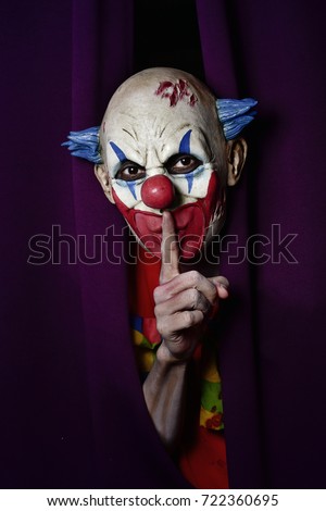 a scary evil clown peering out from a purple stage curtain, with his forefinger in front of his lips, asking for silence