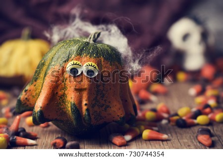 a pumpkin with funny eyes surrounded by halloween candies on a rustic wooden table and a skull in the background