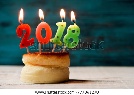 closeup of four lit number-shaped candles of different colors forming the number 2018, as the new year, on a cheesecake on a table, with a blank space on the right