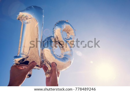 closeup of the hands of a young man holding some silvery number-shaped balloons forming the number 18 against the blue sky