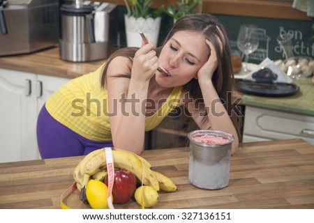 Sad young woman leaning on the kitchen counter at home and eating ice cream. She is too depressed to keep a diet.