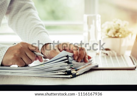 Businessman hands holding pen for working in Stacks of paper files searching information business report papers and piles of unfinished documents achieves on laptop computer desk in modern office