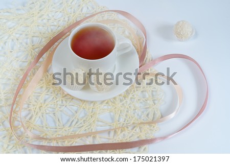 Gentle still life in light tones. Cup of hot tea, Rafaello\'s candy and satin ribbons put in the form of two hearts on a napkin weaved from straw.