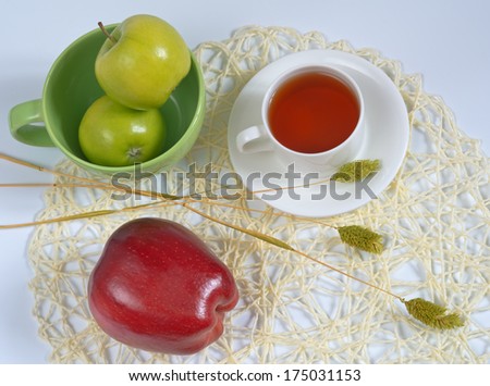 Bright still life in complementary colors - green and red on a white background. Three apples, plate, cup of tea and bouquet from dry blades on a straw napkin.
