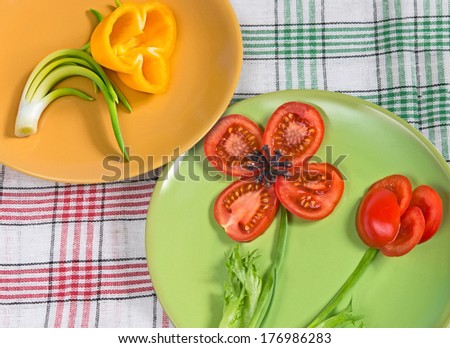 Two painted plates with vegetables laid out on them in form of flowers and checkered napkins.