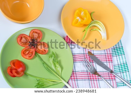 Two painted plates with vegetables laid out on them in form of flowers and checkered napkins.