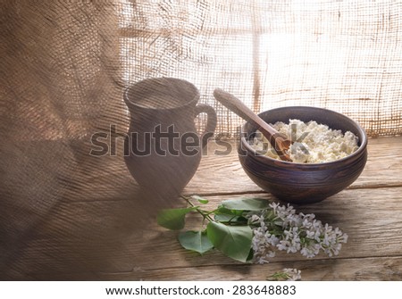 Milk in pitcher and curd in clay bowl and lilac flowers on wooden table next to window and waving in wind curtain.
