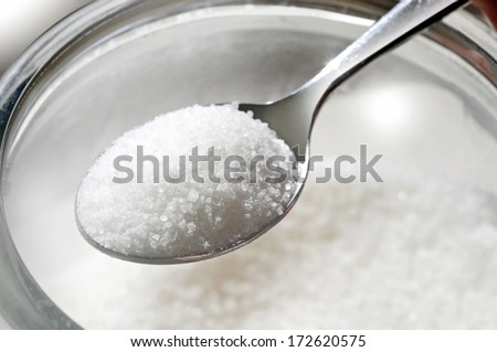 closeup studio photo of spoon full of sugar over glass jar on white background with focus on sugar crystals