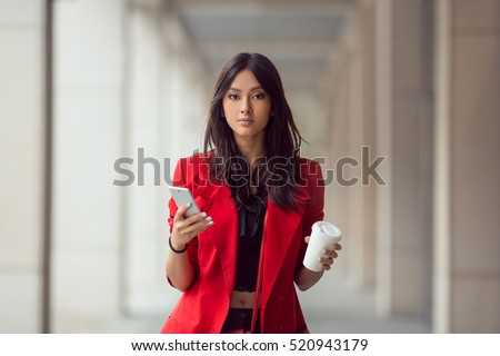 Young Asian woman with smartphone standing against street blurred building background and looking. Fashion business photo of beautiful girl in red casual suite with phone and cup of coffee