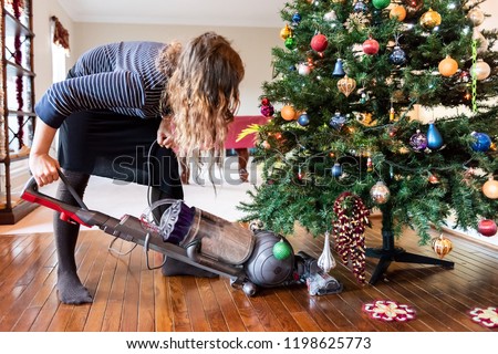 Young woman cleaning with vacuum cleaner, vacuuming under Christmas Tree needles with New Years ornaments on hardwood wooden floor