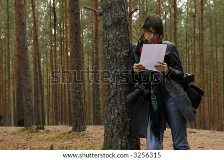 girl with a camera gear is leaning against the tree and reading something from a paper