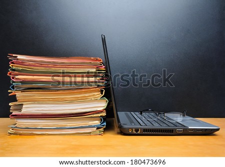 Laptop and office files on wooden table in front of black wall