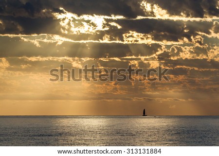 Sailing boat on the horizon with rays of sunlight filtering through clouds onto the calm ocean below.