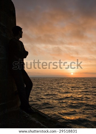 A silhouetted young man watches sunrise from the end of a pier.