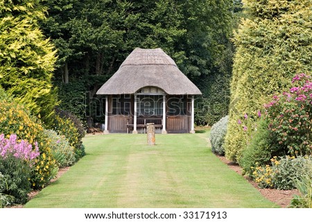 A summerhouse in a domestic garden in summer, with flowers, trees, mowed lawn, and stone sundial