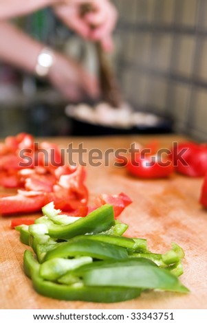 Red and green peppers chopped on a wooden chopping board in a domestic kitchen with cooking in background