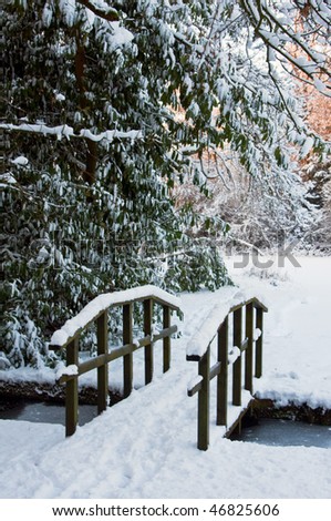 A footbridge covered in snow passes over a frozen offshoot from a lake in winter. Lake is surrounded by woodland, with warm sunset light in the background.